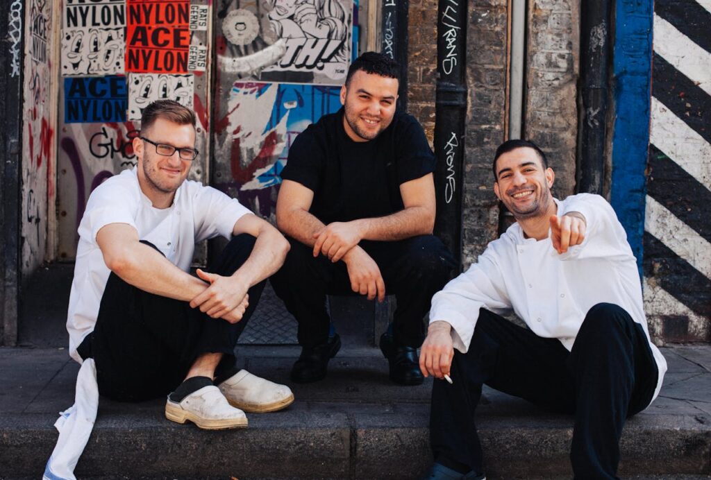 Three men sitting casually in an urban setting with graffiti-covered walls, smiling and wearing casual and chef attire, capturing a relaxed vibe perfect for a Budapest stag do atmosphere.