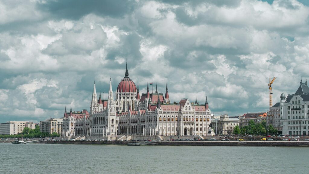 View of the Hungarian Parliament building along the Danube River in Budapest, under a dramatic cloudy sky, showcasing iconic architecture.