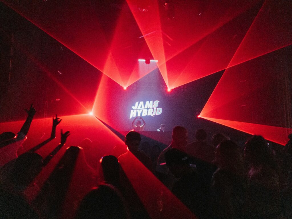 A nightclub in Budapest with a crowd enjoying an energetic DJ set under red laser lights. The 'Jams Hybrid' logo is displayed on the screen, creating a vibrant atmosphere, ideal for a Budapest stag party or a pub crawl Budapest stag experience.