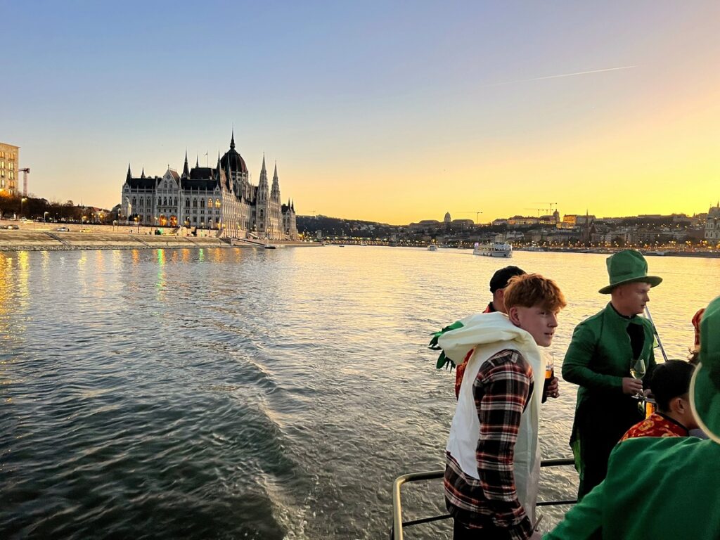 Group of friends dressed in themed outfits enjoying a private riverboat party on the Danube, with the stunning Hungarian Parliament building in the background during sunset