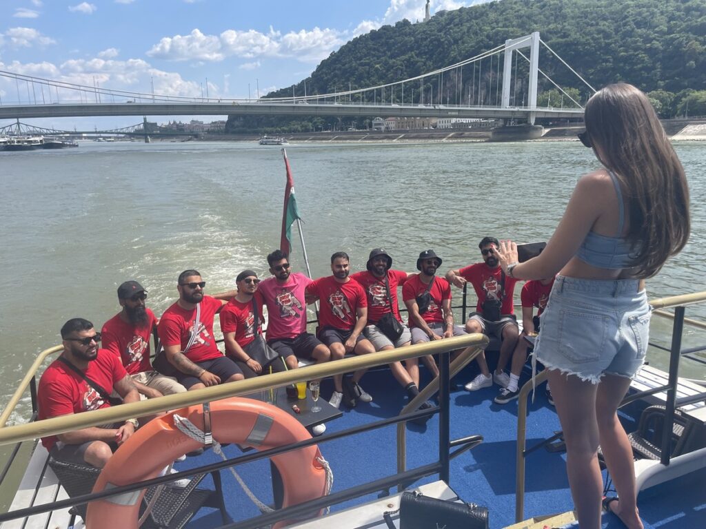 Group of friends on a stag do, wearing matching red T-shirts, posing for a photo on a boat cruising down the Danube River in Budapest. A woman is taking their photo as they enjoy the sunny day, with the Elizabeth Bridge and green hills visible in the background.