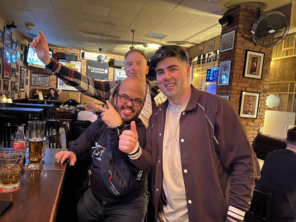 Three friends enjoying a night out in a cozy pub. They’re smiling, giving thumbs up, and posing with glasses of beer on the bar. The pub’s interior is warmly lit, with sports posters and framed photos on the walls, creating a relaxed and friendly atmosphere.