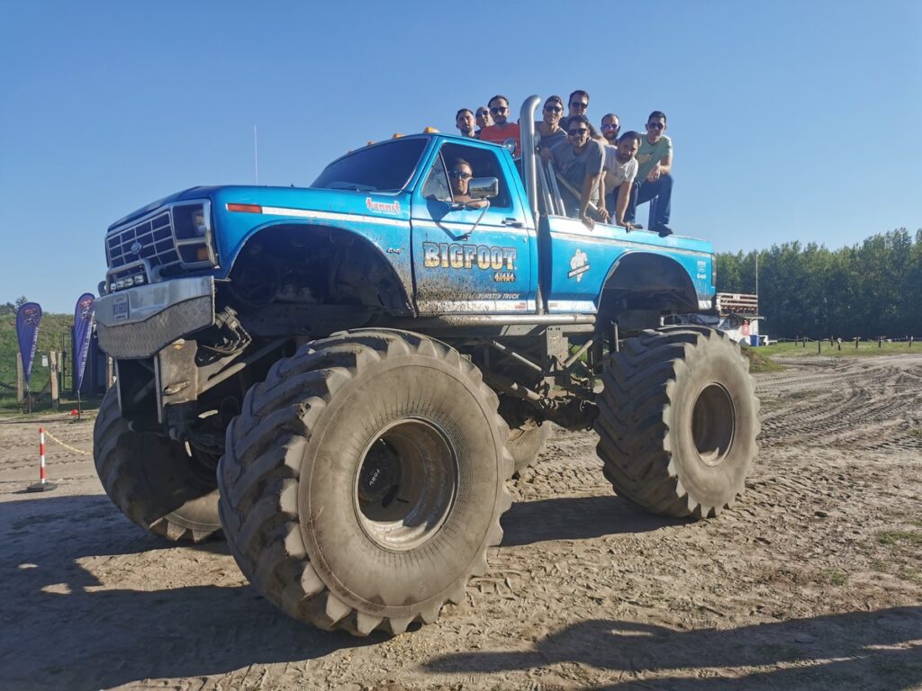 A group of friends enjoying a stag do adventure, standing on top of a large blue monster truck labeled 'Bigfoot' with massive tires. The group is smiling and posing for a photo under a clear blue sky in an open, outdoor setting, creating an exciting and adventurous atmosphere.