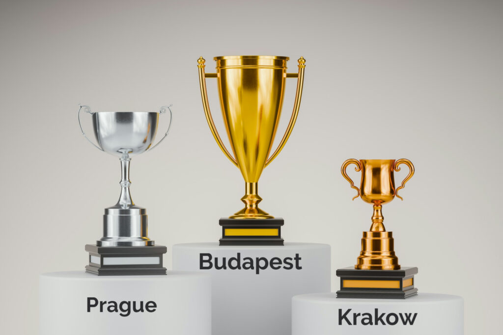 Podium with three trophies representing a comparison between European stag do destinations. A large gold trophy labeled 'Budapest' stands in the center on the highest podium, indicating first place. To the left, a silver trophy labeled 'Prague' is in second place, and to the right, a bronze trophy labeled 'Krakow' is in third. This visually emphasizes Budapest as the top choice for stag dos.