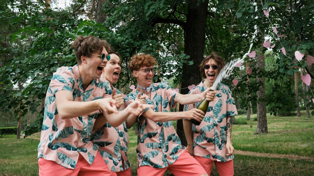 Group of friends celebrating outdoors, dressed in matching tropical-themed outfits with pink shorts and green leaf-patterned shirts. One person is popping a champagne bottle, spraying it with excitement, while everyone laughs and cheers. The scene is set in a green, wooded area, creating a lively and fun atmosphere perfect for a stag do celebration.