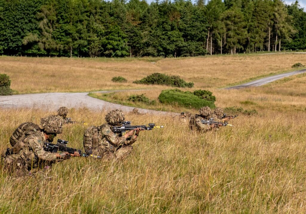 Shooting range challenges with soldiers in camouflage gear participating in tactical field exercises, lying in tall grass with rifles aimed, surrounded by open fields and dense forest in the background.