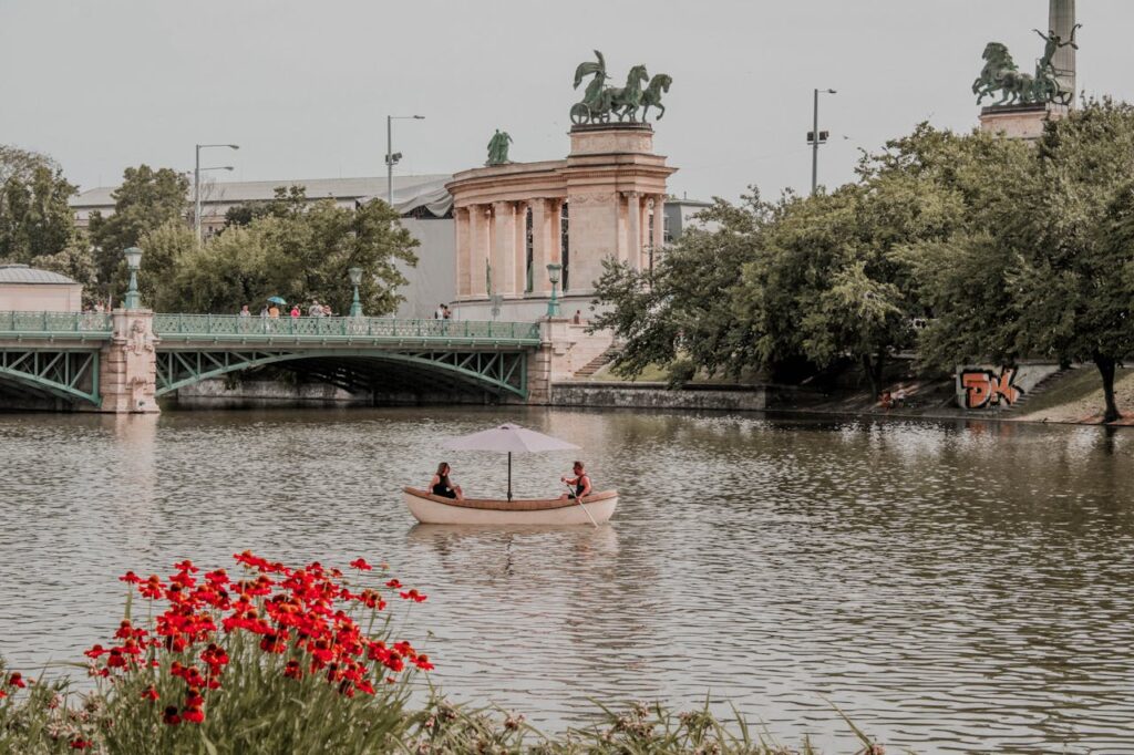 Epic Budapest stag adventures with a summer boat ride on a peaceful lake surrounded by red flowers, lush greenery, and iconic Budapest monuments in the background.