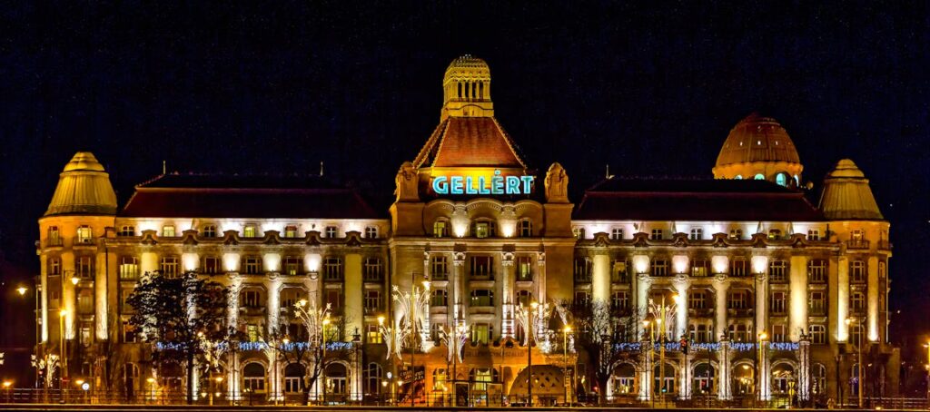 Night view of the illuminated Gellért Hotel and Spa in Budapest, showcasing its iconic architecture with glowing lights against the dark sky, creating a grand and historic atmosphere.