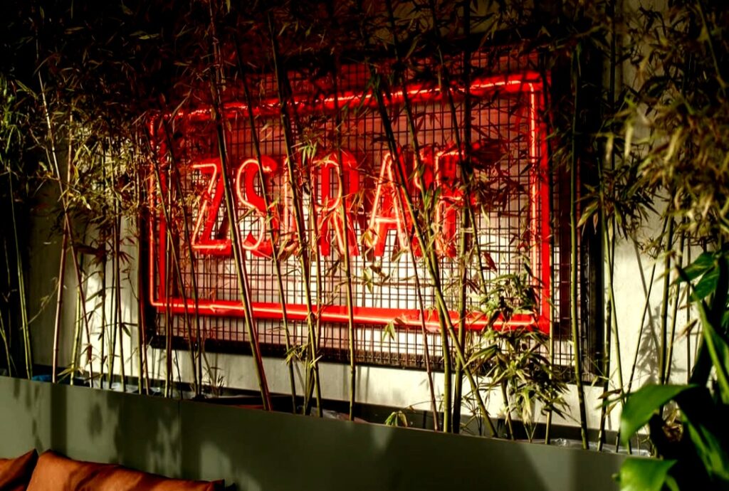 Zsiráf Cocktail Bar's vibrant red neon sign glows behind lush greenery, creating a stylish and cozy ambiance in this popular Budapest bar.