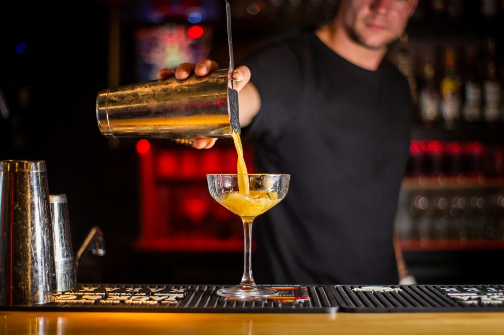 A bartender in a stylish Budapest cocktail bar pours a vibrant drink from a shaker into an elegant coupe glass, set against a dimly lit background with red and gold hues, showcasing the city's thriving mixology scene.