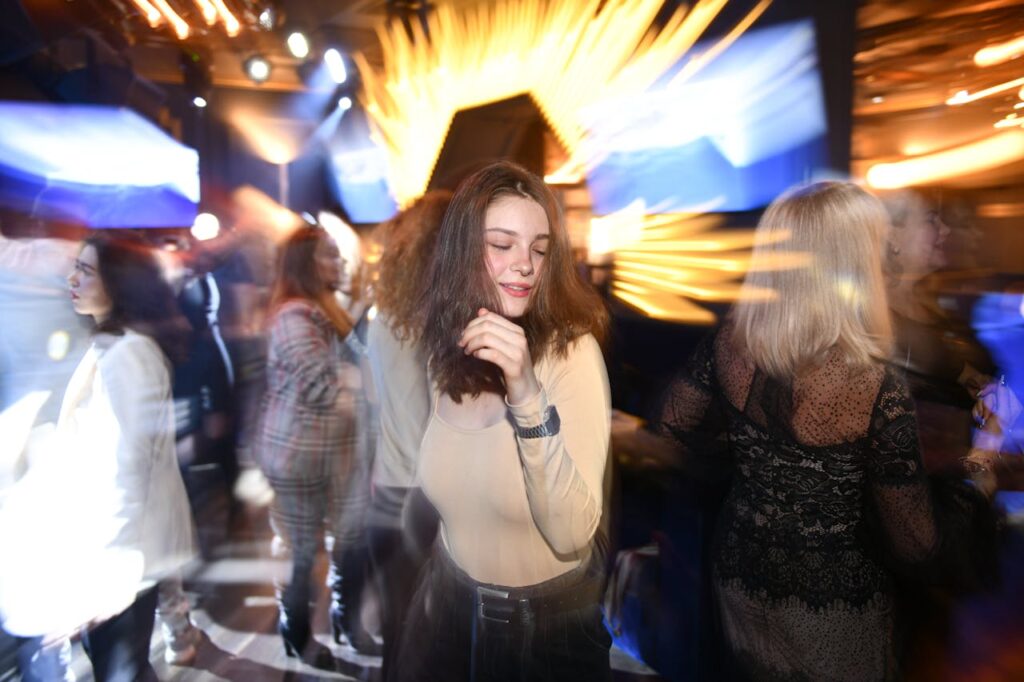 A young woman dancing with a relaxed smile, surrounded by dynamic lights and a lively atmosphere at a nightclub.