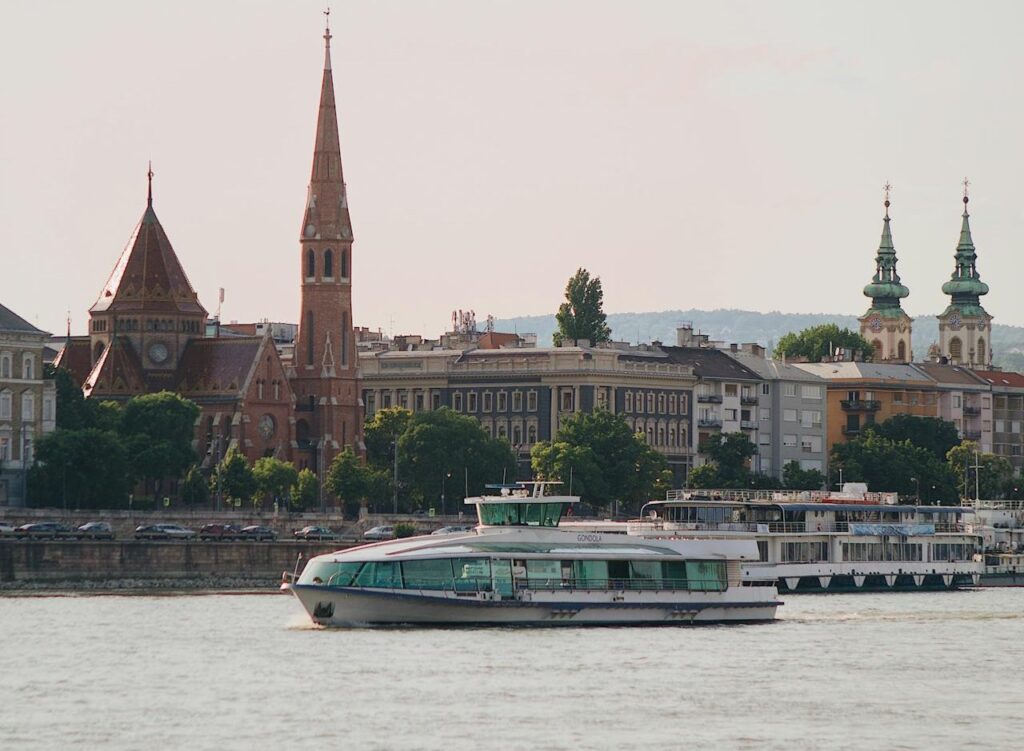 A scenic view of a river cruise boat sailing past historic buildings and churches along the Danube in Budapest