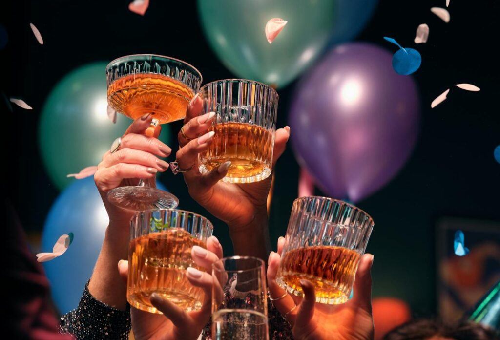 A festive celebration with hands raising glasses of whiskey, surrounded by colorful balloons and falling confetti.