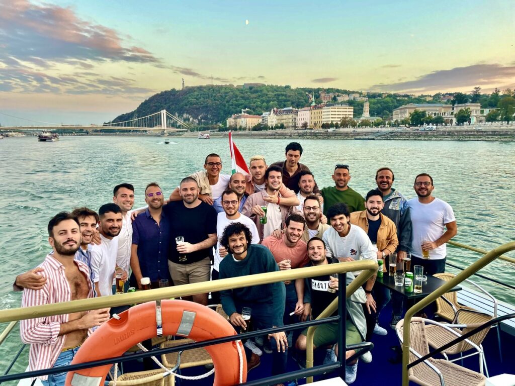 A large group of men enjoying a Budapest stag do on a boat cruise along the Danube River, holding drinks and celebrating against a scenic backdrop of the city and Gellért Hill at sunset. One of the most popular activity according to Budapest stag do reddit threads.