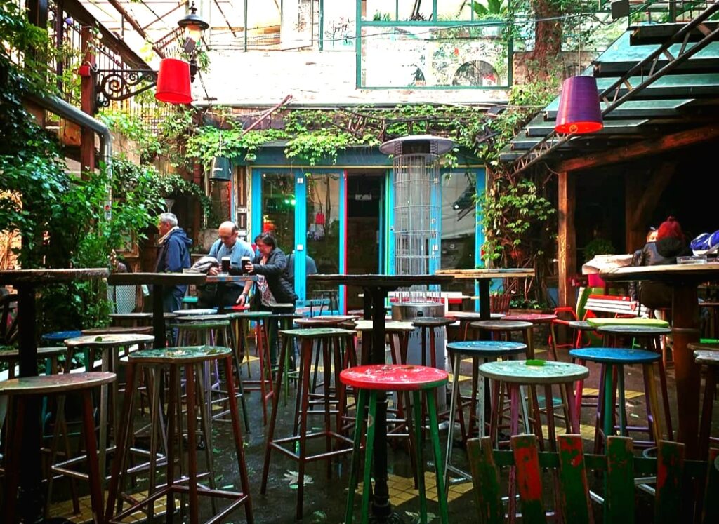 A vibrant ruin bar in Budapest with colorful mismatched stools, rustic wooden tables, and lush greenery, creating a unique and eclectic atmosphere as people enjoy drinks in the background. One of the most popular Budapest stag do reddit threads.