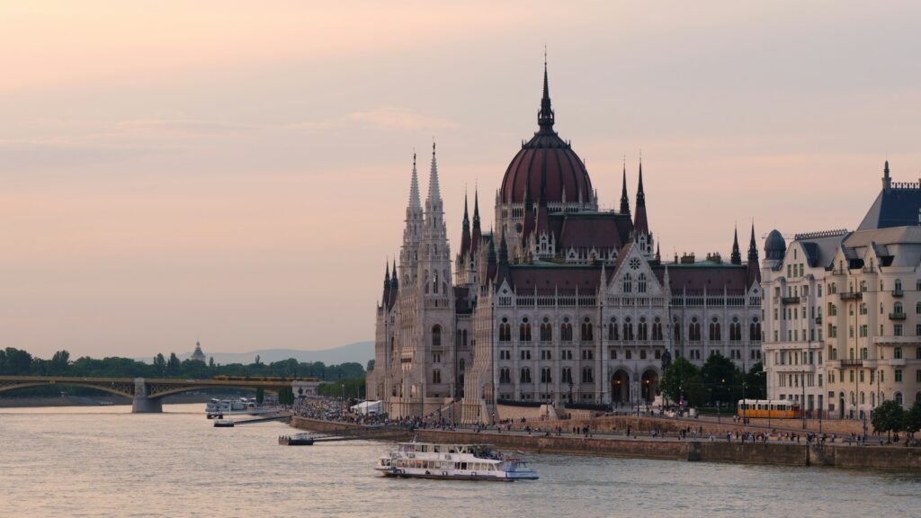 The Hungarian Parliament Building in Budapest at sunset, with a river cruise boat on the Danube, a yellow tram passing by, and crowds along the promenade, a scene often discussed on Budapest stag do Reddit.