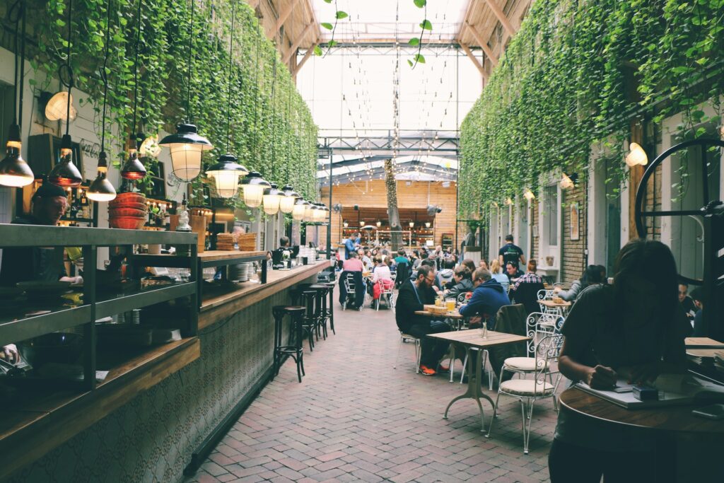 A lively indoor market-style café with hanging greenery, warm lighting, and a mix of rustic and modern decor, where people are dining, socializing, and working in a cozy atmosphere.