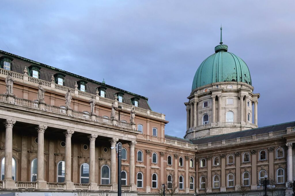 A view of Buda Castle in Budapest, Hungary, showcasing its grand historic architecture with a green dome and classical columns against a cloudy sky.