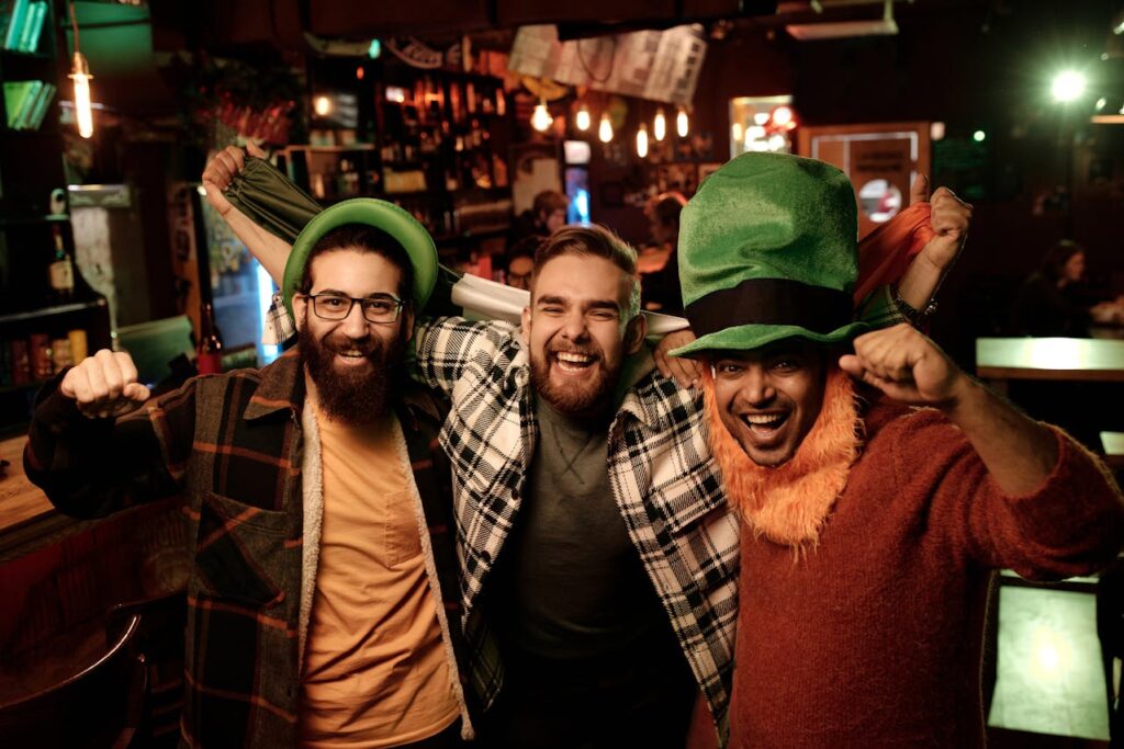 Three men are celebrating in a lively pub, dressed in festive outfits with a green hat and an orange beard, cheering with an Irish flag behind them as part of a Budapest stag do Reddit.