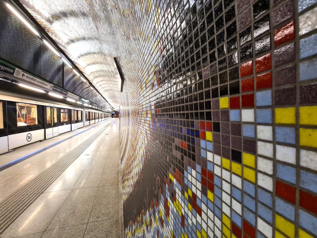 A modern Budapest metro station with a sleek design, featuring a colorful mosaic-tiled wall, bright lighting, and a waiting train on the platform, creating a dynamic urban atmosphere.