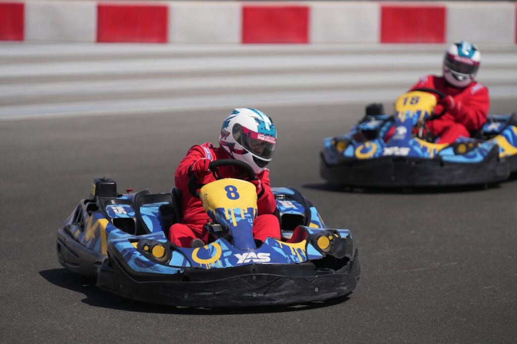 Two go-kart racers in red racing suits and helmets compete on an outdoor track, driving blue and yellow karts with determined focus.