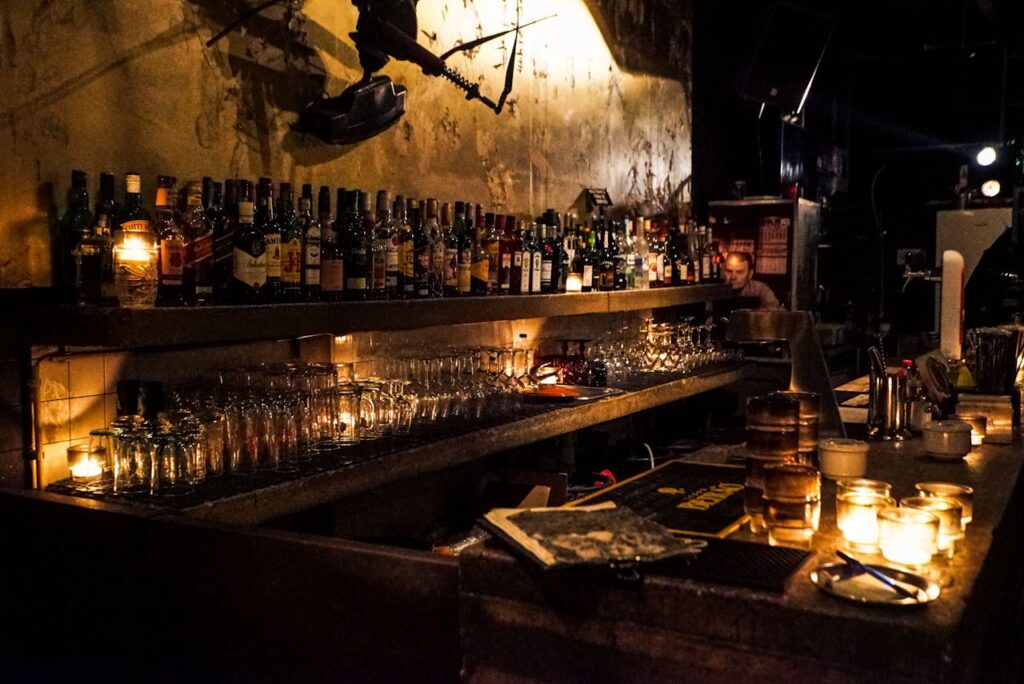 A dimly lit, atmospheric bar with a long counter lined with liquor bottles and glassware, illuminated by candlelight, creating a warm and cozy ambiance.