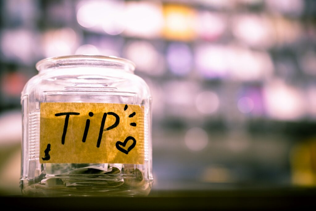 A glass tip jar with a handwritten label that says 'TIP' and a heart symbol, containing cash and coins, placed on a counter with a blurred background of lights.